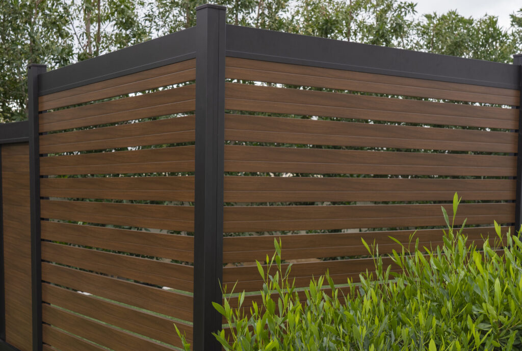 Modern aluminum fence with horizontal brown wood-grain slats and black posts installed beside green plants.